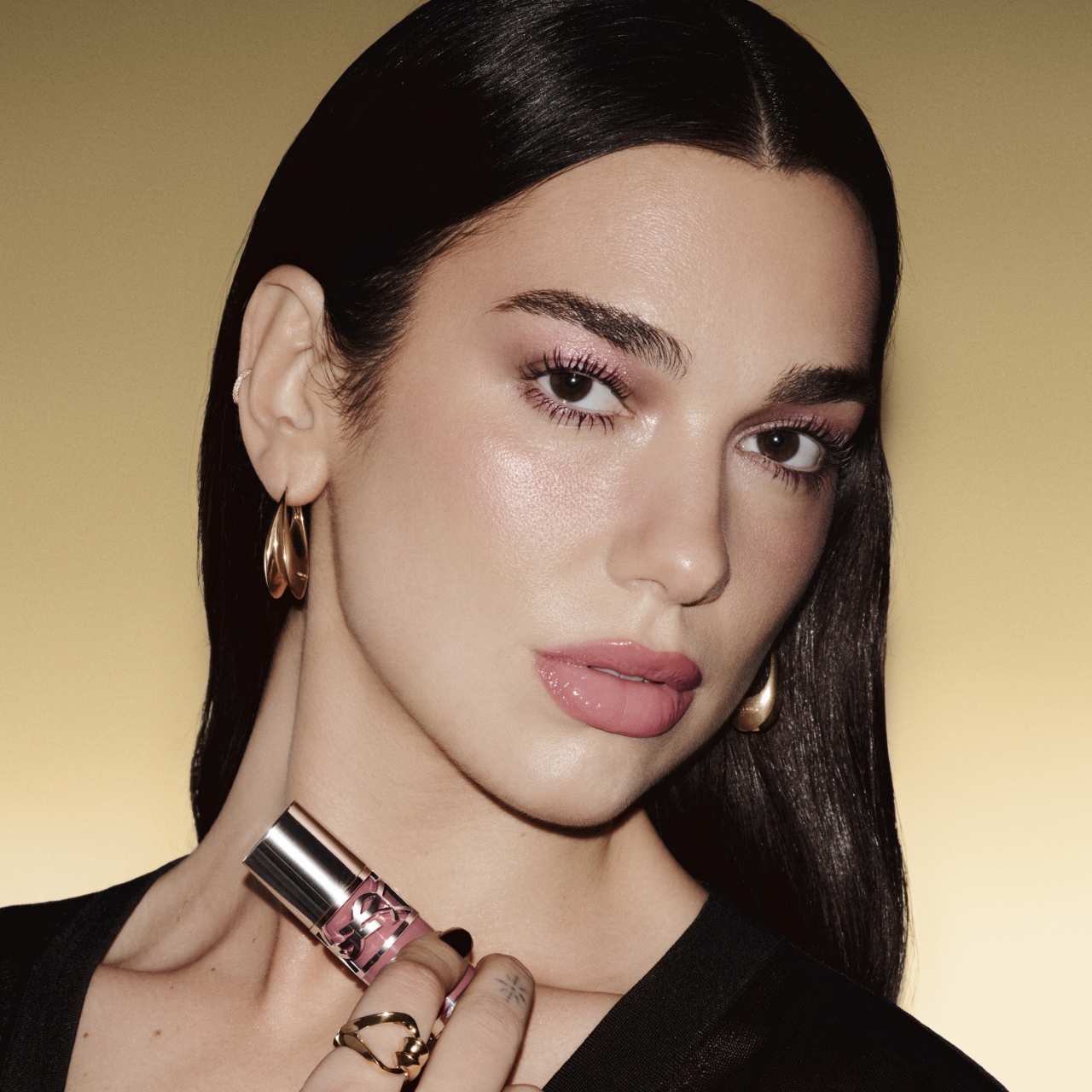 Dua Lipa, con lunghi capelli scuri e un trucco rosa tenue, tiene un rossetto YSL vicino alla guancia, guardando direttamente lo spettatore su uno sfondo dorato.