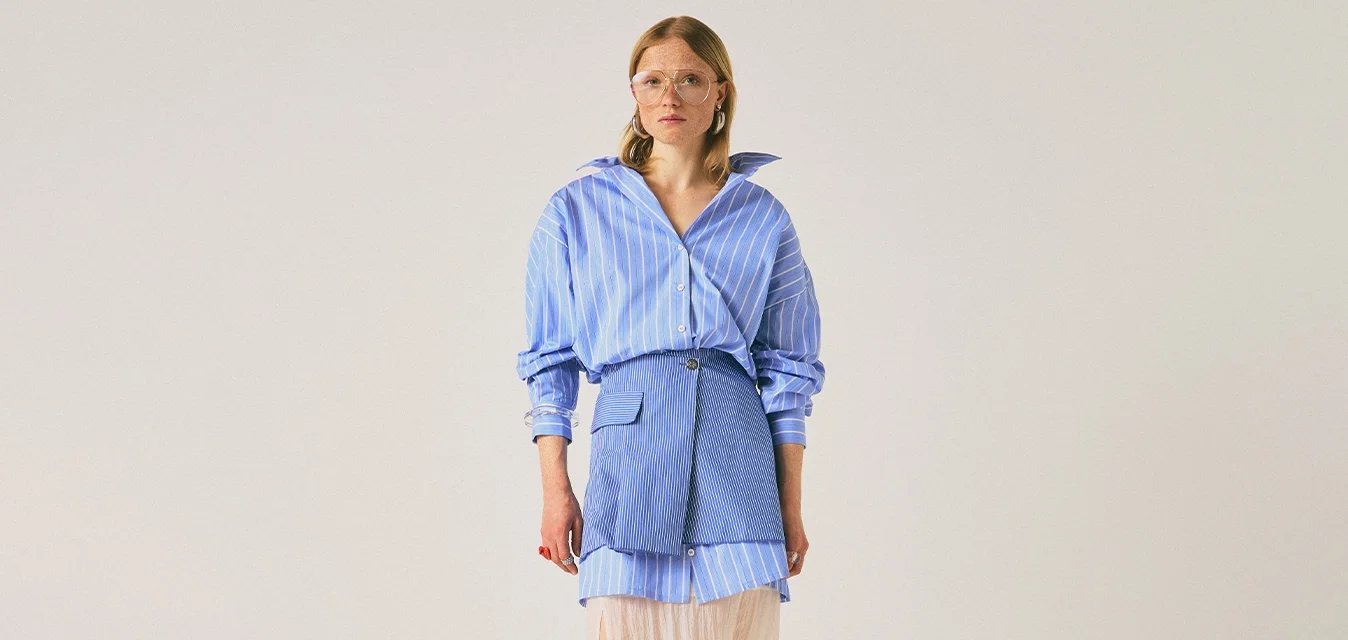 Donna che indossa una camicia a righe blu oversize, una mini gonna blu con tasca, una gonna midi trasparente beige, tacchi bianchi e occhiali grandi, in piedi contro uno sfondo neutro.
