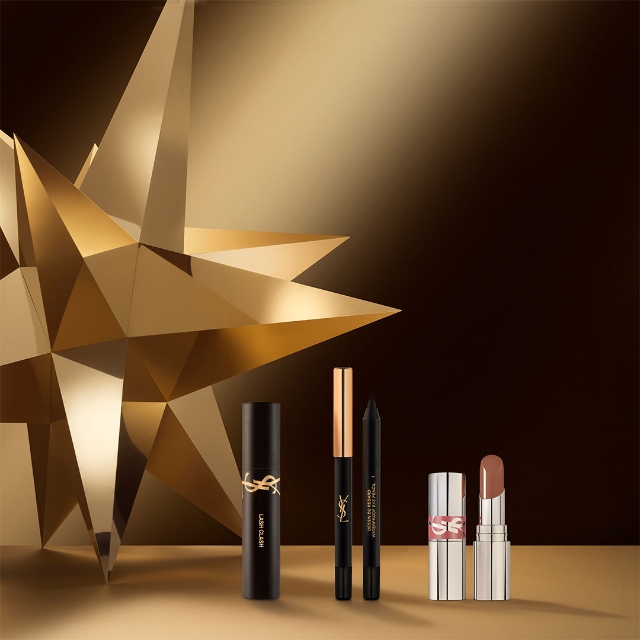 Un mascara YSL Lash Clash, un eyeliner nero e un rossetto nude sono artisticamente disposti davanti a una grande stella geometrica dorata su uno sfondo scuro e sfumato.