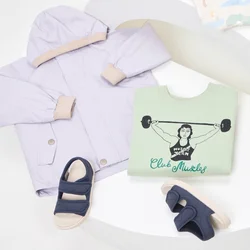 Giacca lilla, felpa grafica verde, camicia a fantasia, jeans azzurri chiari e sandali con accenti colorati disposti su una vetrina curva bianca.