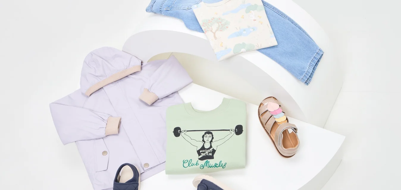 Giacca lilla, felpa grafica verde, camicia a fantasia, jeans azzurri chiari e sandali con accenti colorati disposti su una vetrina curva bianca.