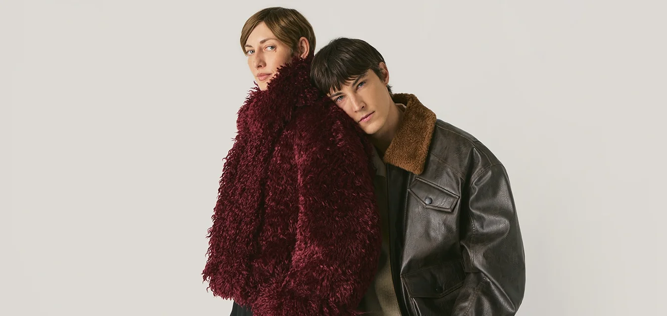 Due giovani adulti che indossano capi esterni dalle texture particolari; uno in un cappotto peloso borgogna, l'altro in una giacca di pelle marrone scuro con colletto in shearling.