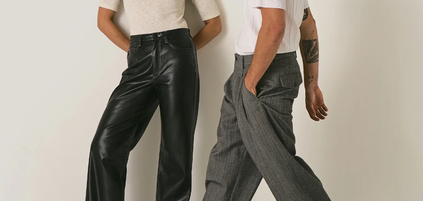 Due adulti alla moda si trovano davanti a un semplice muro beige; donna in camicia bianca e pantaloni neri, uomo in camicia bianca e pantaloni larghi grigi che cammina.