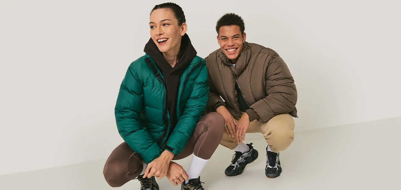 Due giovani adulti accovacciati e sorridenti, indossando giacche casual invernali e scarpe da ginnastica, su uno sfondo grigio chiaro uniforme.