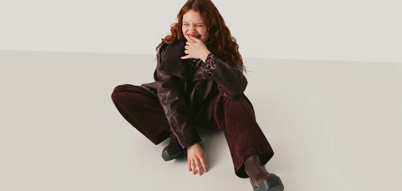 Giovane donna con capelli ricci rossi seduta a terra con pantaloni e giacca viola scuro, sorridente e che tocca il viso su sfondo neutro.