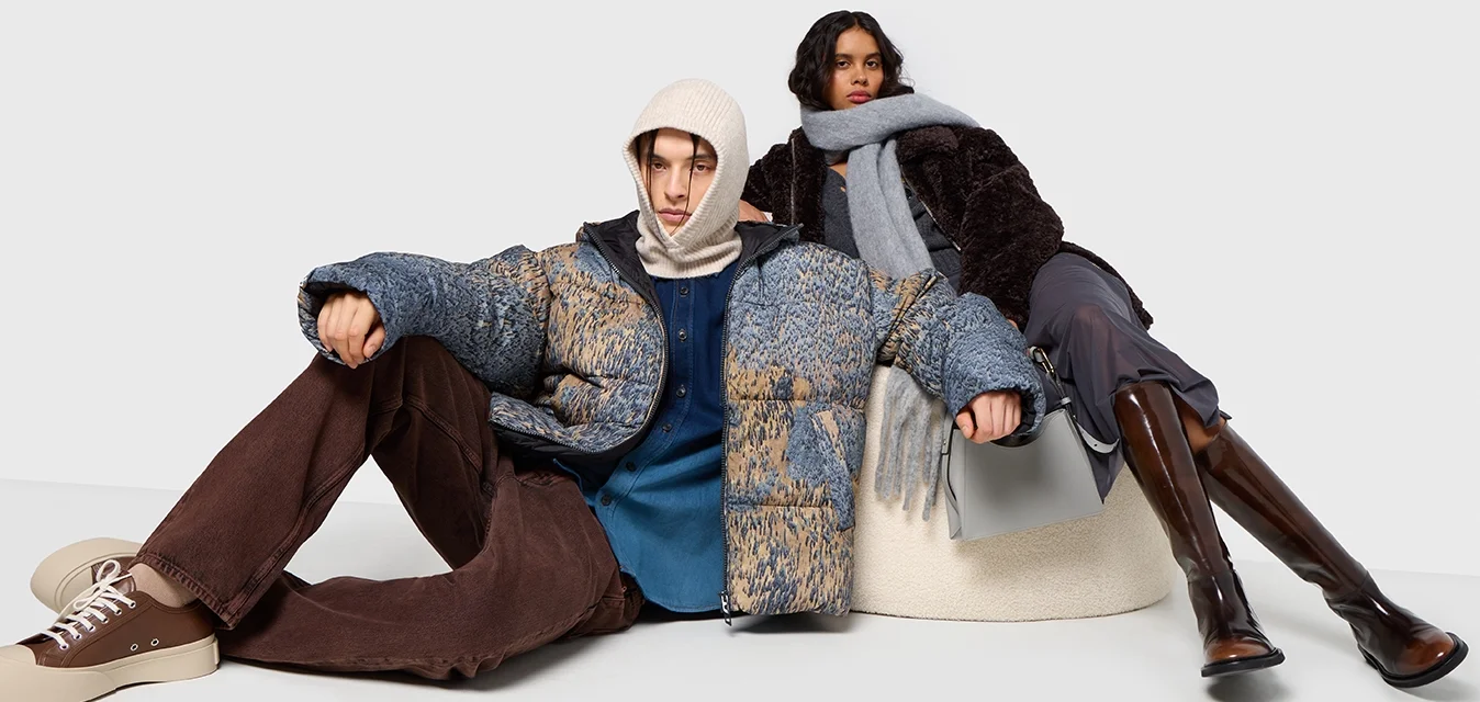 Giacca imbottita blu e beige, sciarpa grigia, pantaloni a campana marroni e stivali marroni. Tessuti morbidi e abbigliamento a strati in una posa seduta.