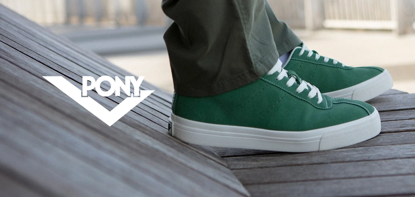 Sneakers in pelle scamosciata verde con suole in gomma bianca, design con lacci e dettagli perforati, abbinate a pantaloni verde oliva su una superficie in legno.