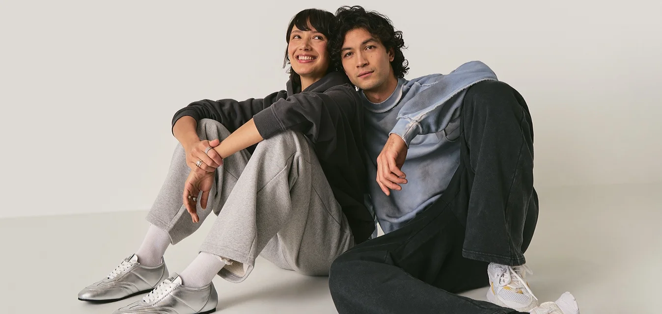 Giovane uomo e donna seduti sul pavimento appoggiati l'uno all'altro, vestiti con abbigliamento casual e sneakers, sfondo neutro.