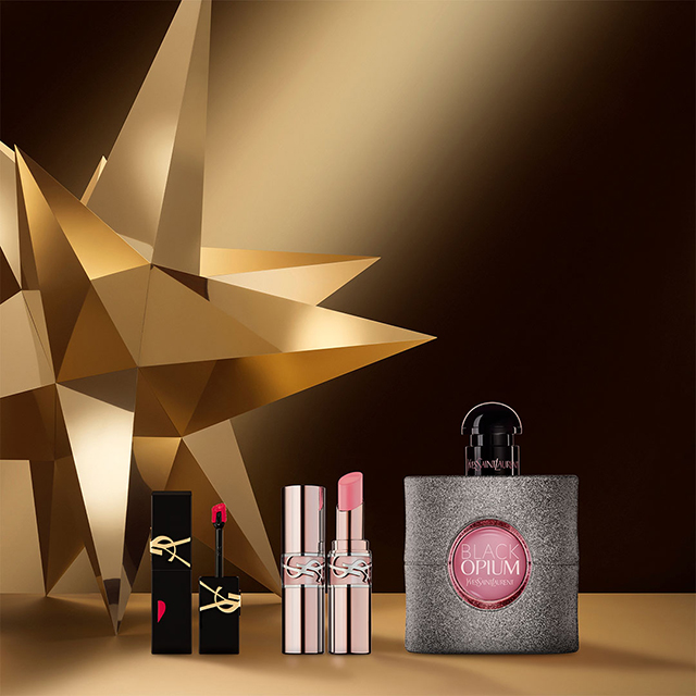 Un lucidalabbra rosso acceso, due rossetti rosa e un flacone di profumo YSL Black Opium sono elegantemente disposti davanti a una stella geometrica dorata scintillante su uno sfondo scuro sfumato.