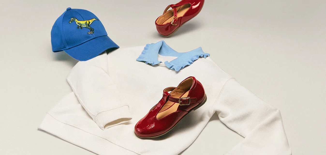 Camicia bianca a maniche lunghe con colletto blu, cappellino blu con dinosauro e un paio di scarpe Mary Jane rosse disposti su una superficie chiara.