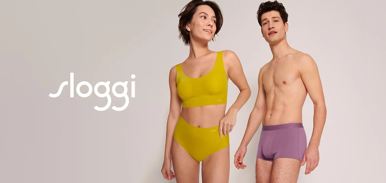 Bra sportivo giallo e culotte alte abbinati a boxer viola, tutti in tessuto liscio con cuciture minime, in piedi su uno sfondo chiaro.