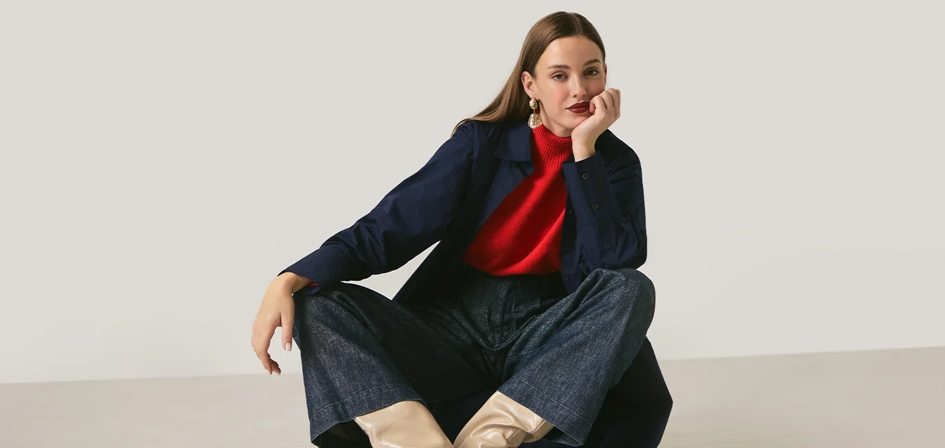 Donna seduta per terra con le gambe divaricate, indossa un maglione rosso, un cappotto blu navy, pantaloni scuri e stivali beige su sfondo chiaro.