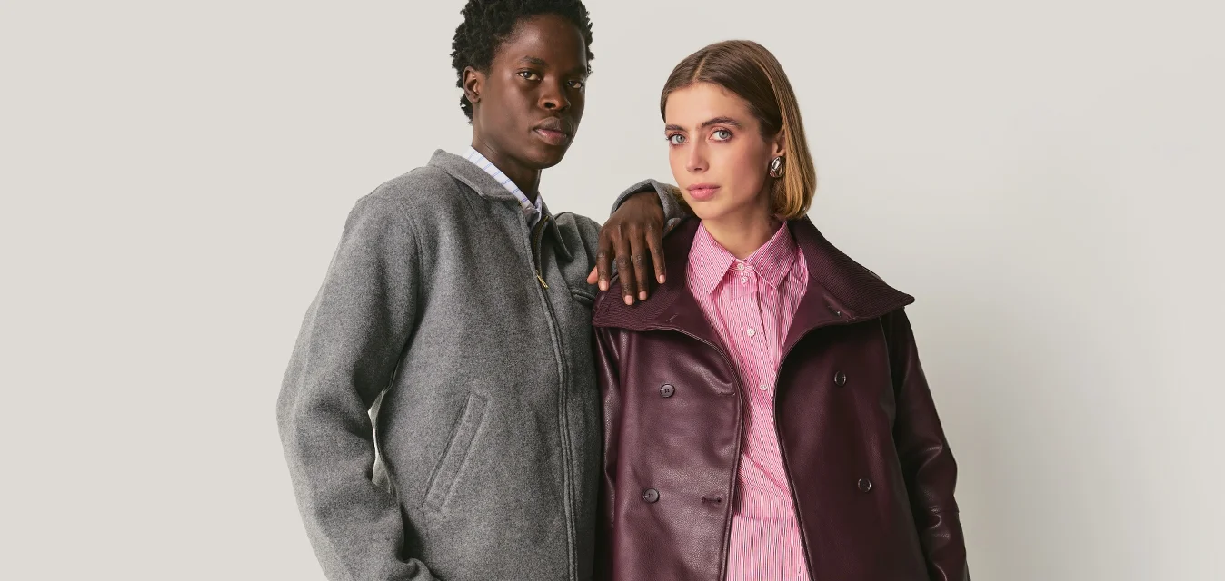 Due persone stanno vicine; una indossa una giacca e pantaloni grigi, l'altra una giacca di pelle viola, una camicia rosa, pantaloni corti e stivali.