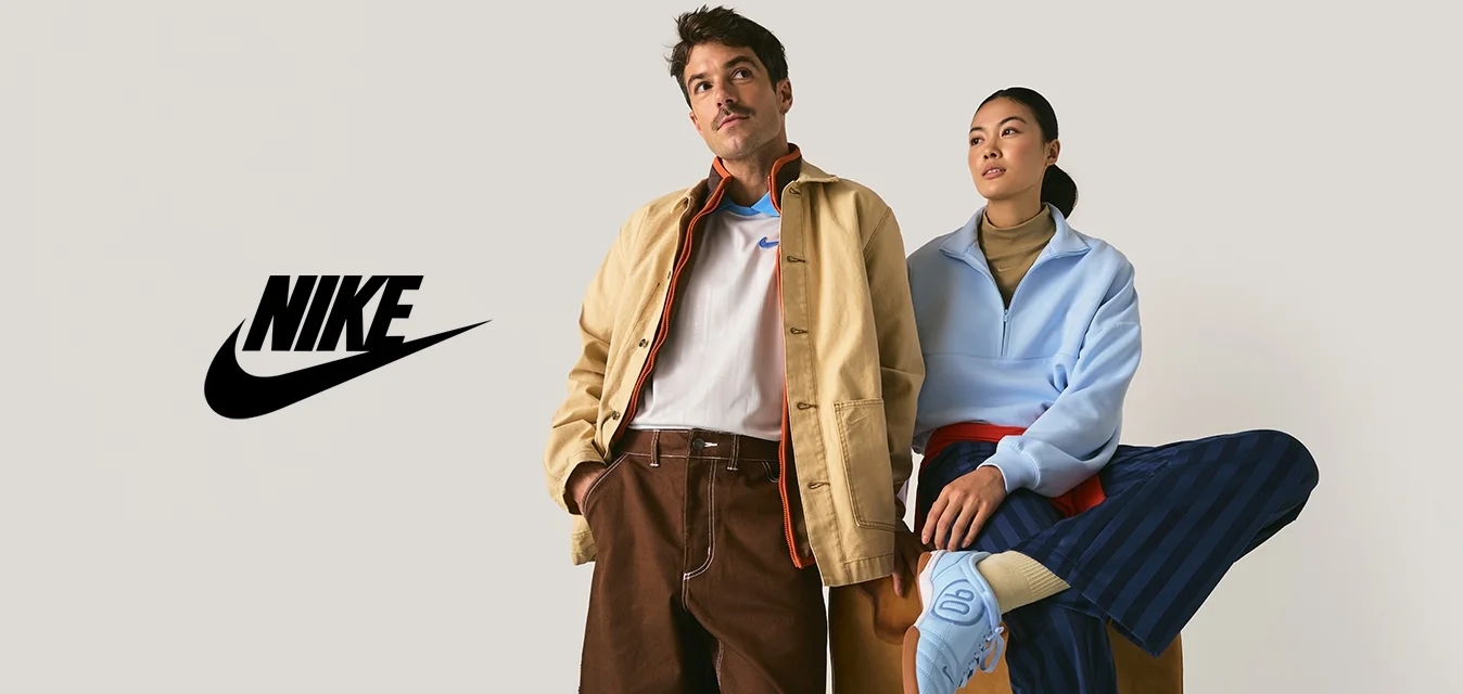 Uomo con giacca beige e pantaloni marroni sta accanto a una donna con pullover azzurro chiaro e pantaloni a righe blu scuro seduta su una panchina.