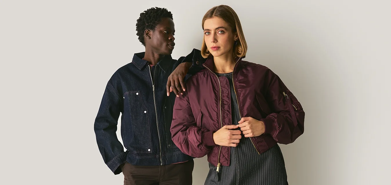 Due modelle posano insieme. Una indossa una giacca di jeans scuro e pantaloni marroni; l'altra sfoggia una giacca bomber borgogna sopra un vestito a righe.