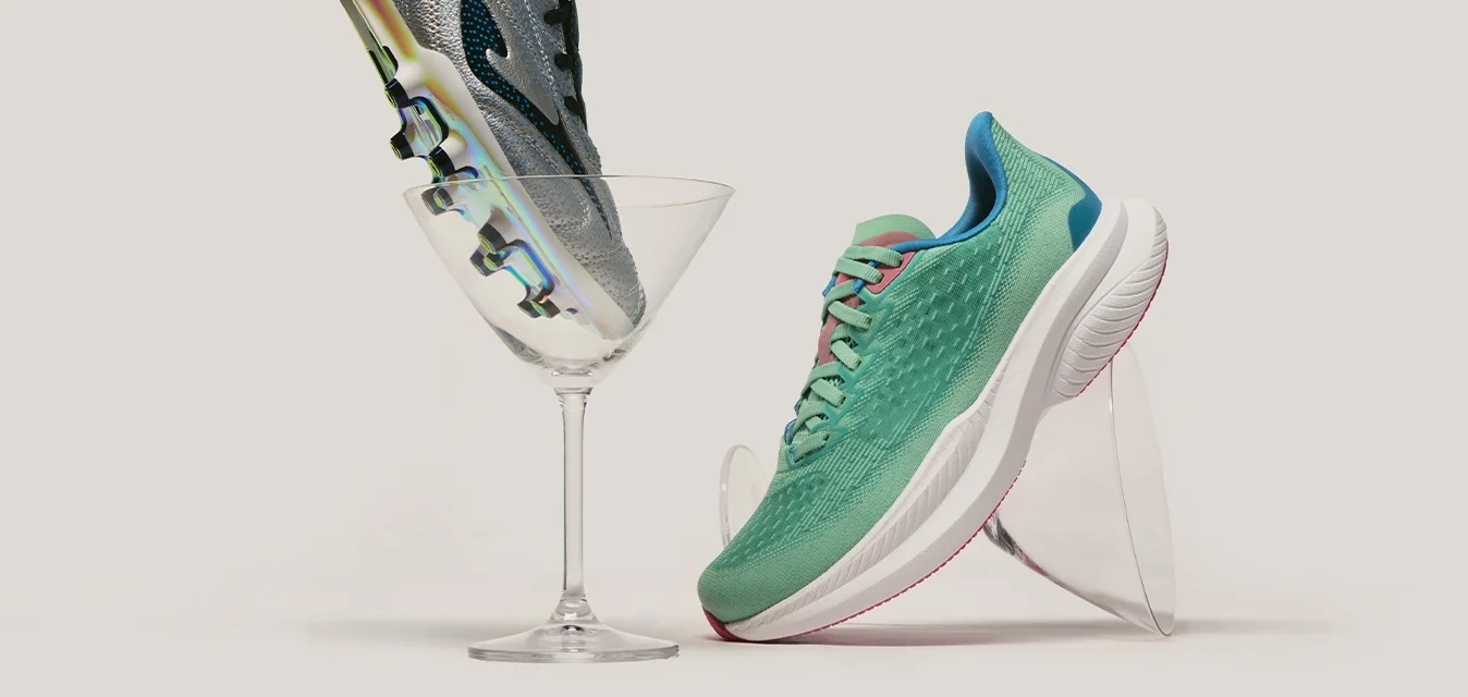 Scarpa da calcio argento equilibrata in un bicchiere da martini accanto a una scarpa da corsa verde inclinata su un supporto trasparente su uno sfondo semplice.