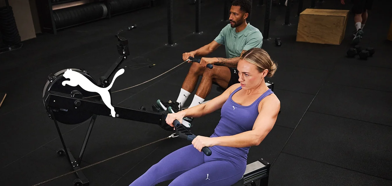 Macchina da remare impostata in una palestra. Due utenti in abbigliamento sportivo; uno indossa leggings viola e una canottiera, l'altro una maglietta verde. Pavimentazione nera e attrezzatura.