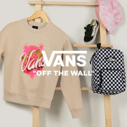 Felpa beige Vans, zaino a scacchi bianco e nero, cappello a secchiello rosa con fantasia e scarpe da neonato nere esposti su una scala di legno.