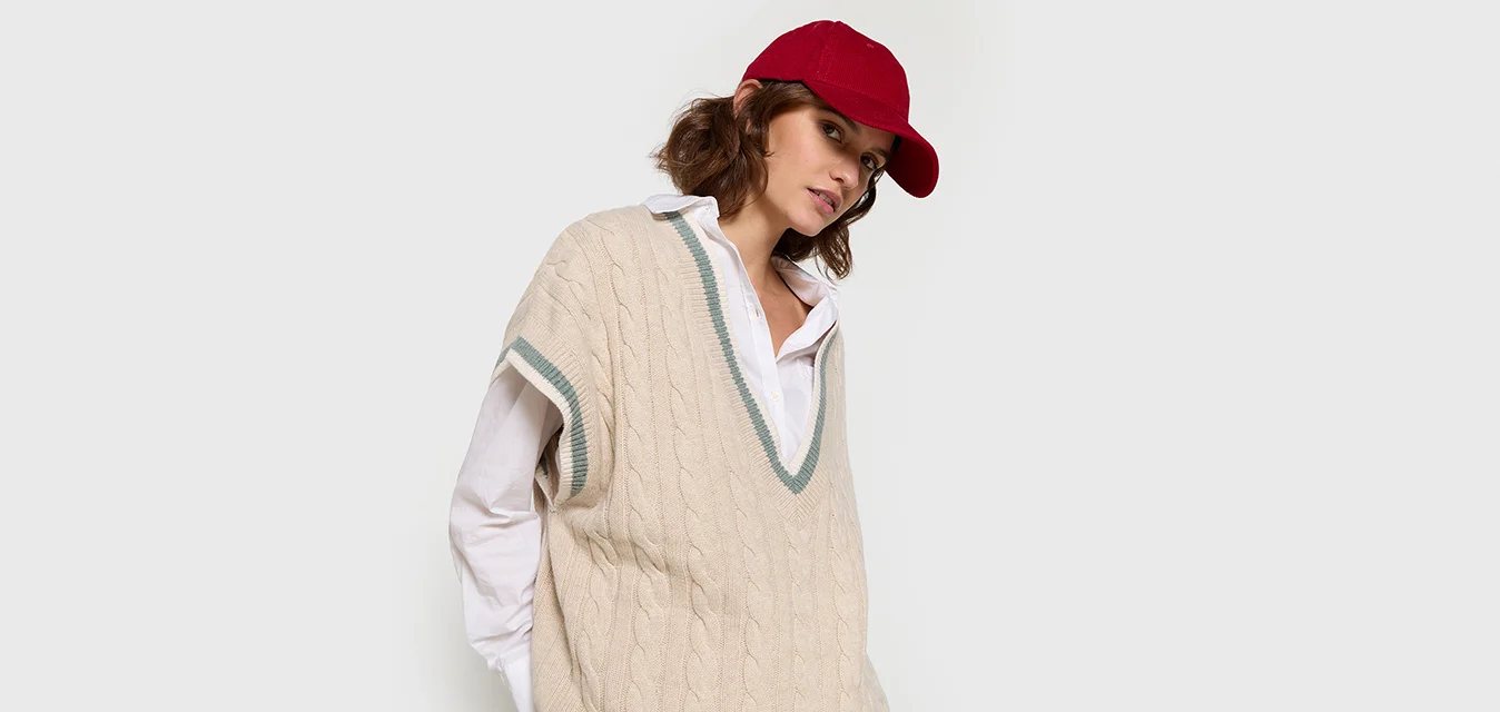 Gilet oversize in maglia cable-knit beige con scollo a V, rifinito con bordi verdi, indossato sopra una camicia bianca con colletto, abbinato a un cappellino da baseball rosso.