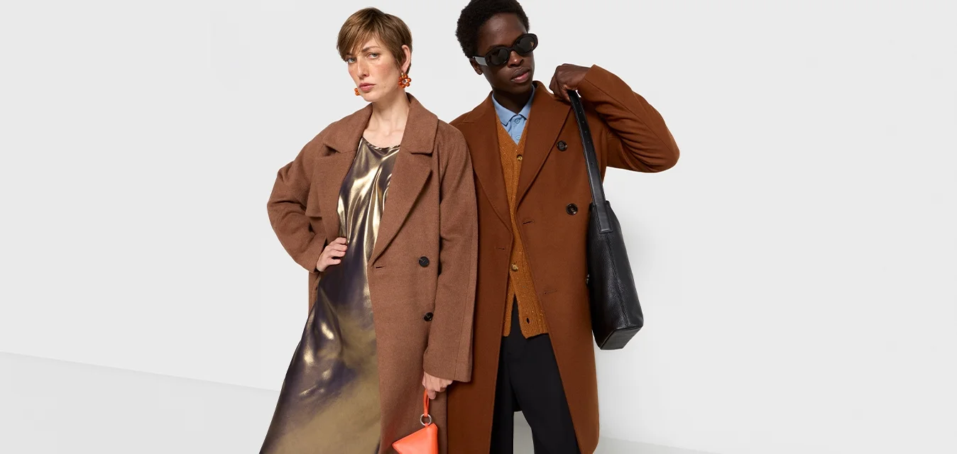 Due persone stanno fianco a fianco. Una indossa un cappotto marrone sopra un vestito metallico, l'altra un cappotto oversize marrone con una camicia blu e pantaloni neri.