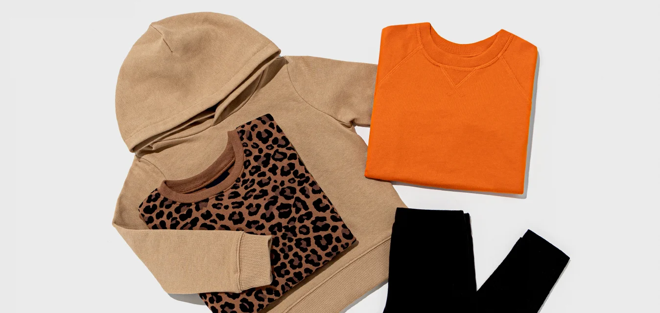 Felpa beige con cappuccio e stampa leopardata, maglione arancione girocollo e leggings neri disposti su uno sfondo grigio chiaro.