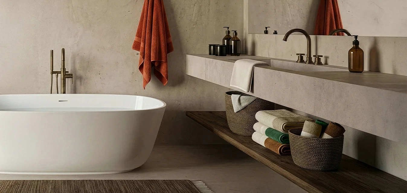 Vasca da bagno indipendente bianca, rubinetto in bronzo, parete in cemento grigio chiaro, mensola in legno con cesti intrecciati, asciugamani colorati e asciugamano arancione appeso.