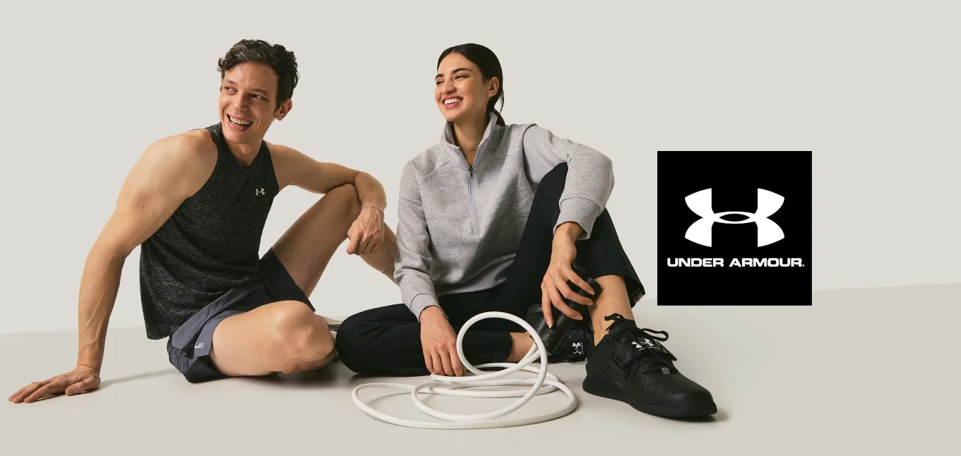 Due adulti in abbigliamento sportivo seduti per terra che sorridono, con una corda da esercizio bianca arrotolata tra di loro su uno sfondo neutro.