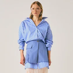 Donna che indossa una camicia a righe blu oversize, una mini gonna blu con tasca, una gonna midi trasparente beige, tacchi bianchi e occhiali grandi, in piedi contro uno sfondo neutro.