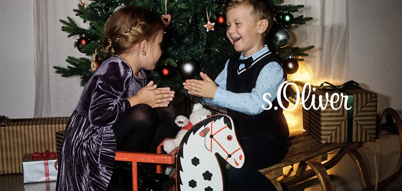 Dei bambini giocano insieme vicino a un albero di Natale. Una ragazza con un vestito di velluto viola è seduta su un cavallo a dondolo rosso; un ragazzo vestito di blu e navy applaude.
