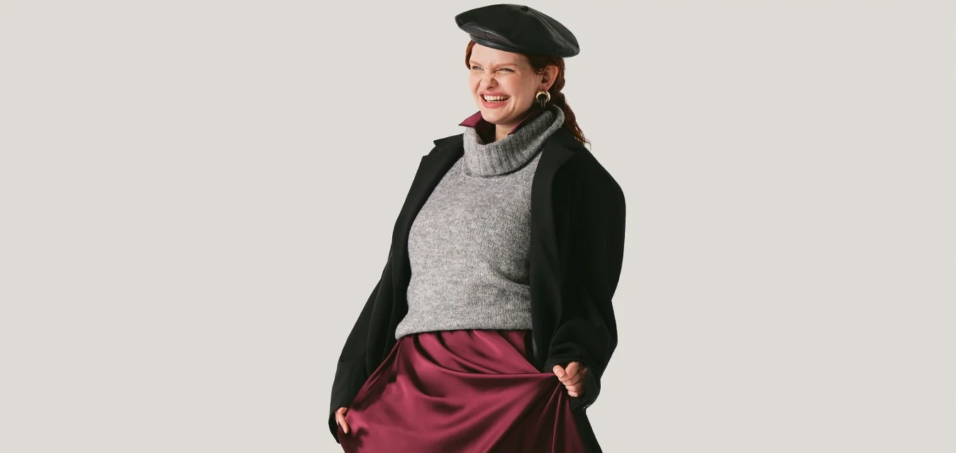 Donna che indossa un basco nero, un maglione grigio, un cappotto nero, una gonna color vinaccia e scarpe nere, camminando con un sorriso davanti a uno sfondo semplice.