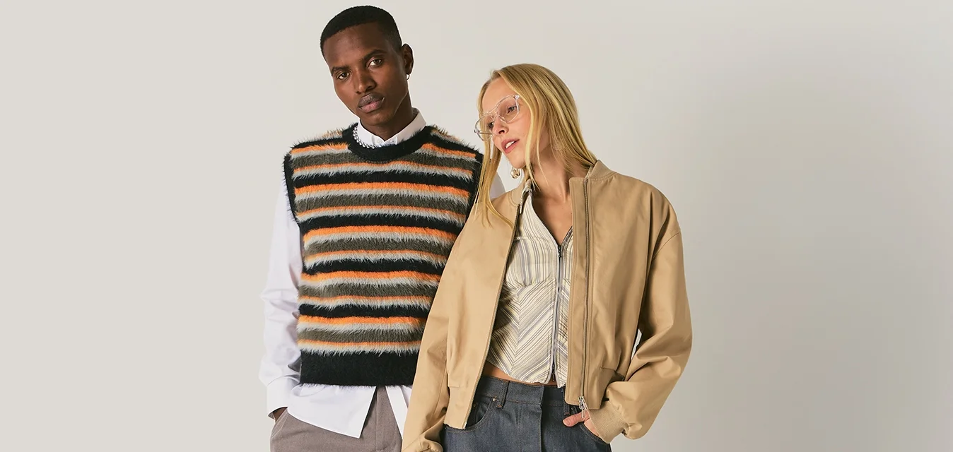 Due modelle in piedi su uno sfondo neutro, una indossa un gilet a righe lavorato a maglia e pantaloni larghi, l'altra una giacca color sabbia, una gonna a fantasia e stivali.