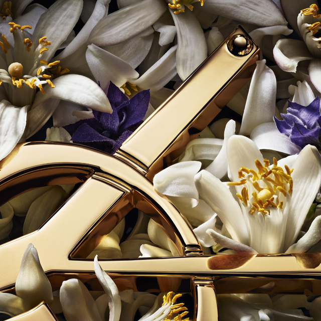 Un primo piano dettagliato mostra l'elegante logo YSL dorato annidato tra un assortimento di profumati fiori bianchi e viola, evidenziati dai loro stami gialli.