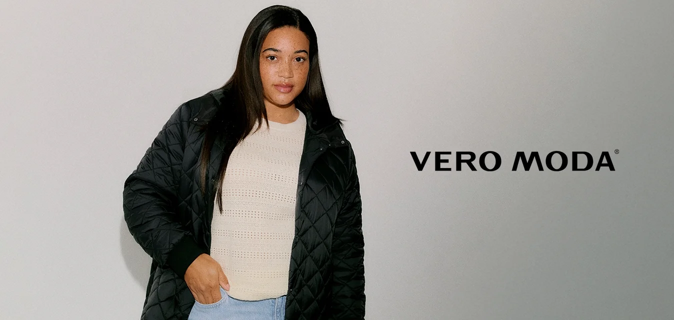 Cappotto lungo nero trapuntato con tasche laterali, polsini a coste e chiusura con cerniera, indossato sopra un top lavorato a maglia color crema e jeans azzurri.