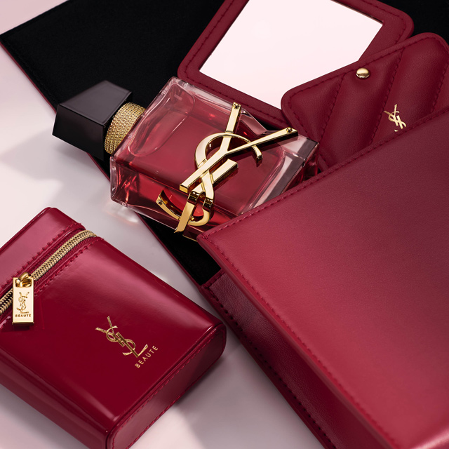 Un flacone di profumo YSL Libre con liquido rosso &egrave; esposto tra lussuosi accessori in pelle rossa, inclusi un compatto con specchio e una pochette con cerniera.
