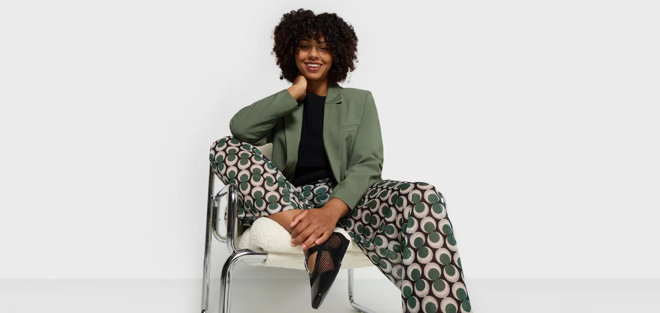 Blazer verde, top nero, pantaloni a gamba larga a motivo con cerchi crema e verdi, e scarpe con dettagli in rete nera. Seduto su una sedia in cromo.