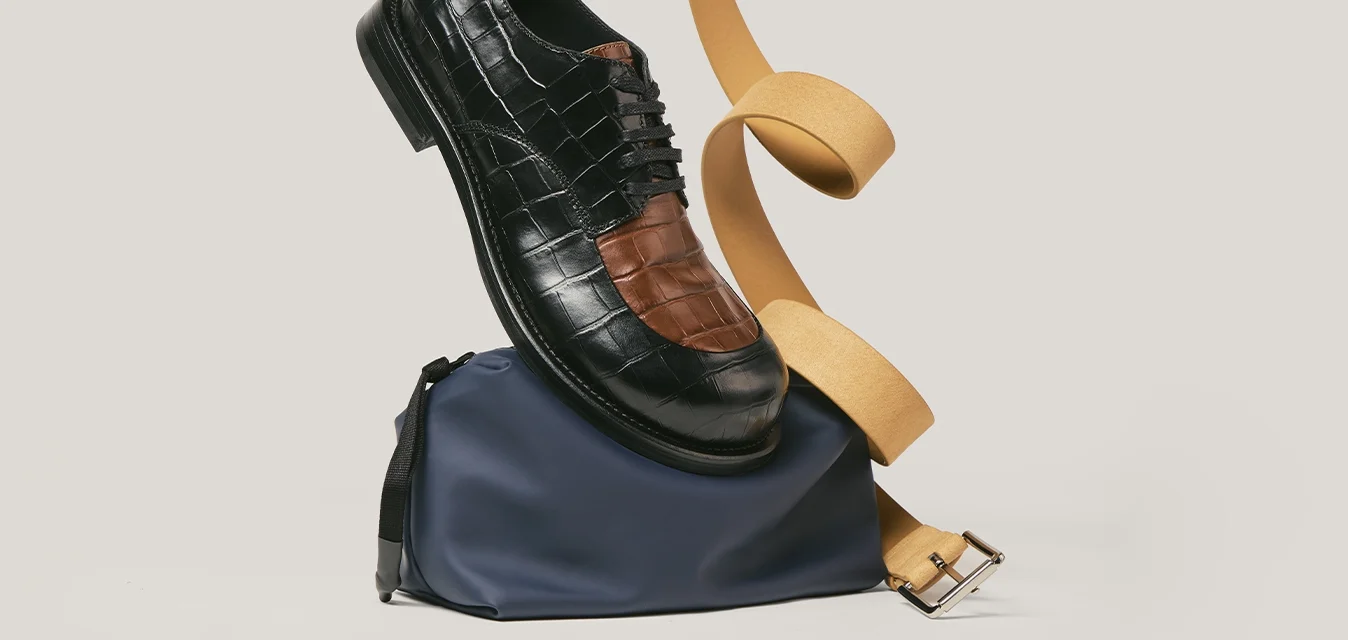 Scarpa elegante in pelle nera e marrone bilanciata su una pochette blu navy con cintura beige arrotolata contro uno sfondo chiaro uniforme.