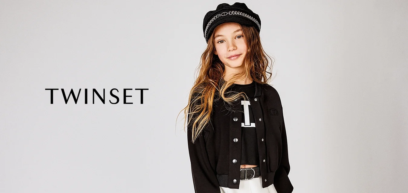 Giacca nera cropped con bottoni a pressione, indossata sopra una camicia nera con una lettera bianca. Gonna bianca svasata e un cappello nero con dettagli in catena.