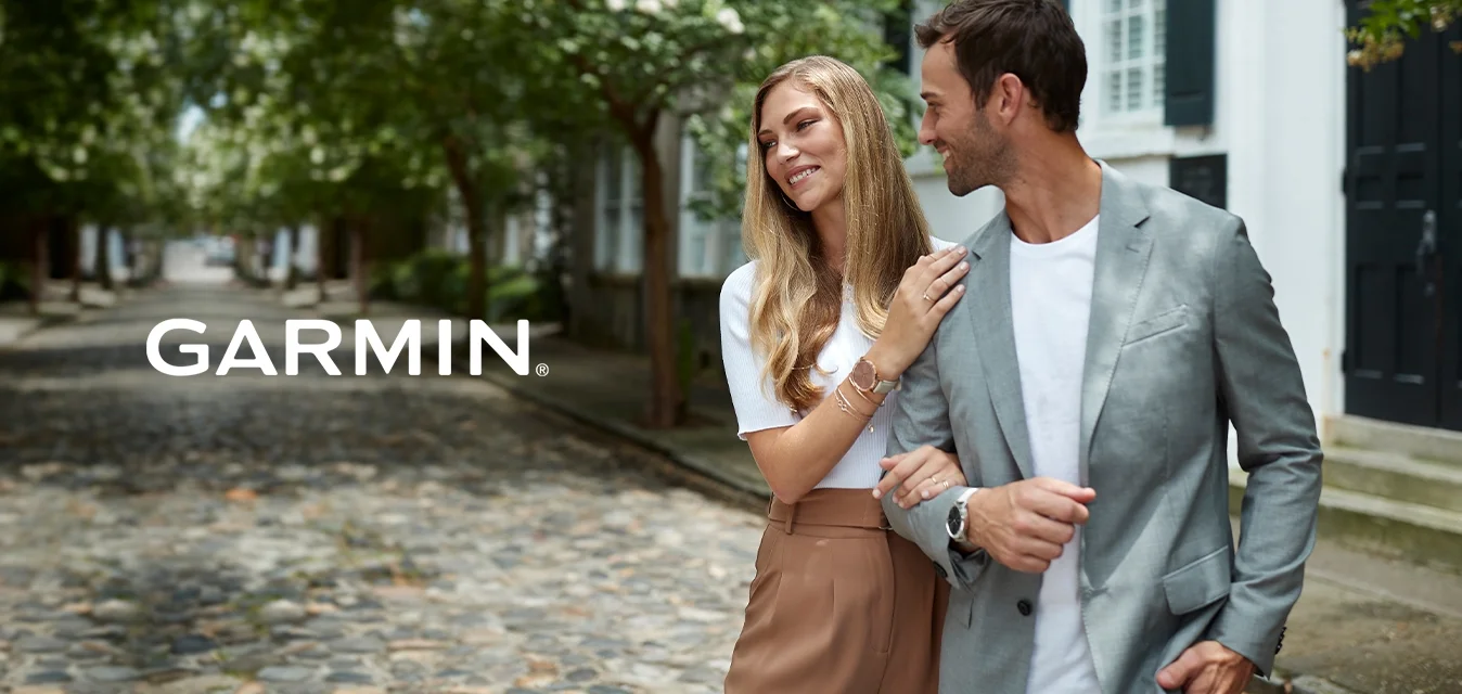 Coppia che cammina su una strada di ciottoli. Donna con un maglione bianco e una gonna beige, uomo con un blazer grigio sopra una maglietta bianca, alberi che fiancheggiano il sentiero.