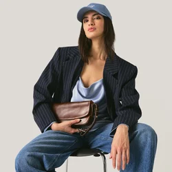 Donna con berretto blu, blazer blu scuro, top in raso, jeans con risvolti ampi, mule bianche con tacco, seduta su una sedia con una pochette marrone in mano.