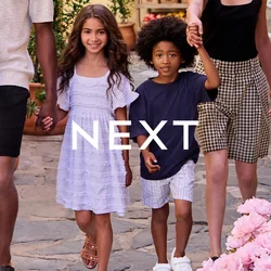 Una famiglia che cammina lungo una strada in acciottolato. L'uomo indossa un maglione leggero e pantaloni corti; i bambini hanno indumenti con motivi vari. Sono presenti piante fiorite.