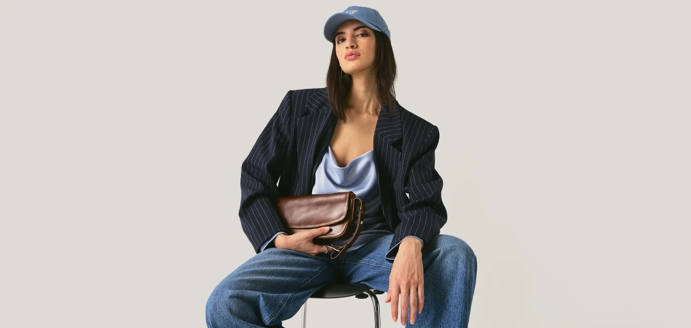Donna con berretto blu, blazer blu scuro, top in raso, jeans con risvolti ampi, mule bianche con tacco, seduta su una sedia con una pochette marrone in mano.