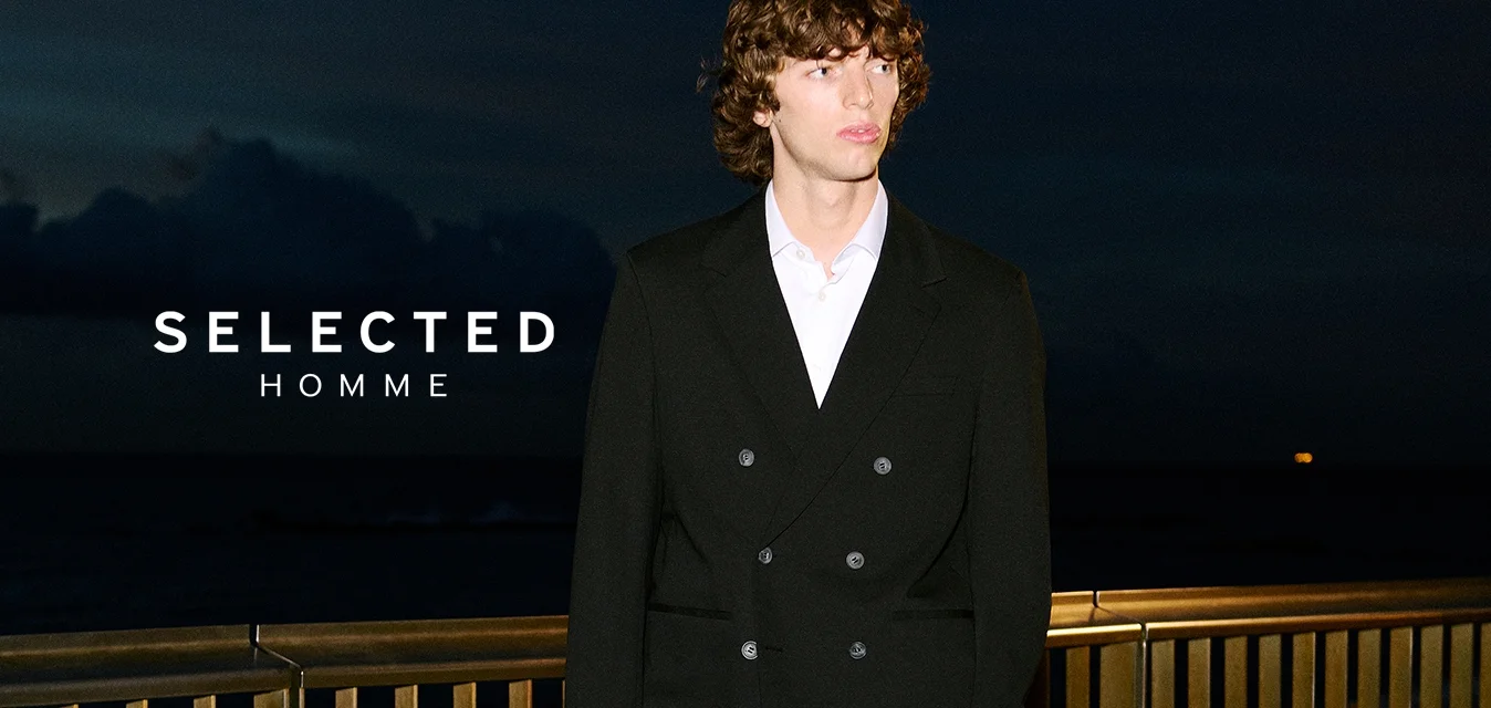 Giovane uomo in un completo nero a doppio petto e camicia bianca che sta in piedi su una terrazza di pietra vicino a un parapetto metallico di notte.