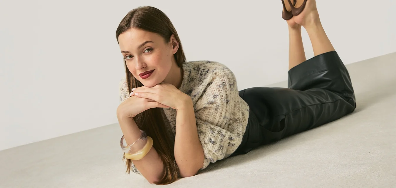 Donna sdraiata sul pavimento con il mento appoggiato sulle mani, indossa un maglione a motivo, pantaloni neri, tacchi marroni e braccialetti, su sfondo semplice.