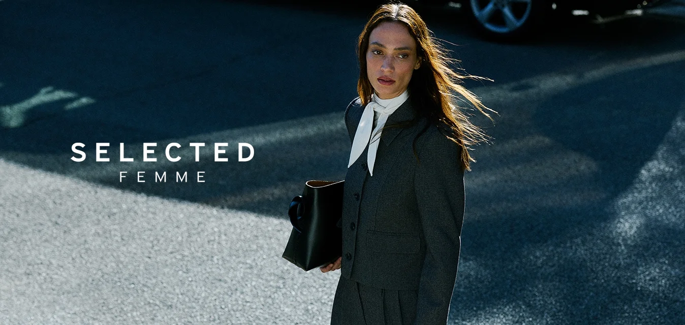 Donna in abito scuro e sciarpa bianca che tiene una borsa nera, mentre cammina su una strada soleggiata vicino a un palo verde e bianco e a un'auto parcheggiata.