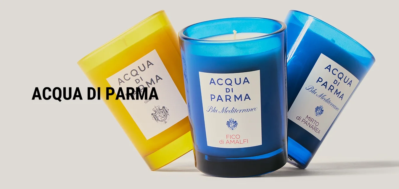 Tre candele profumate in contenitori di vetro giallo e blu etichettati Acqua di Parma, disposte inclinate su uno sfondo grigio chiaro.