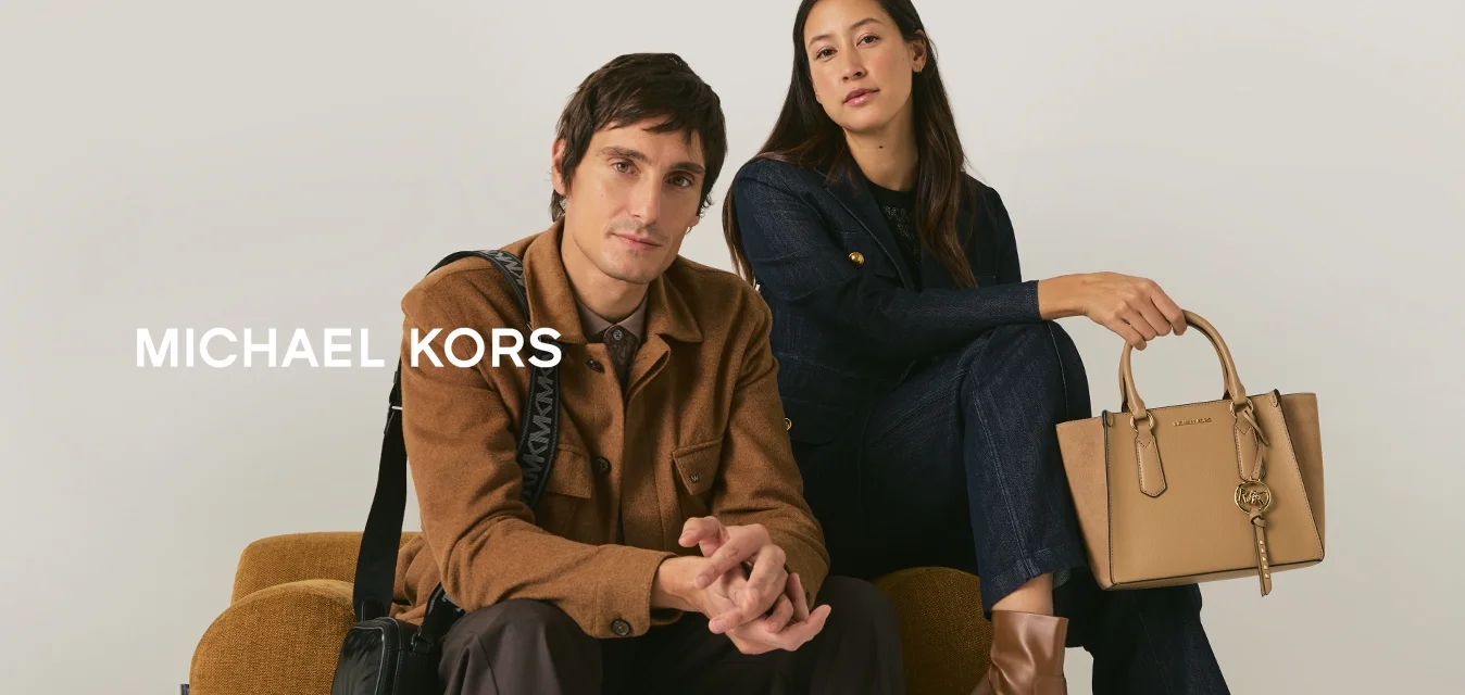 Uomo e donna seduti su un divano di tessuto color senape; l'uomo indossa una giacca marrone con una borsa nera a tracolla, la donna indossa un completo scuro con una borsa beige e stivali.