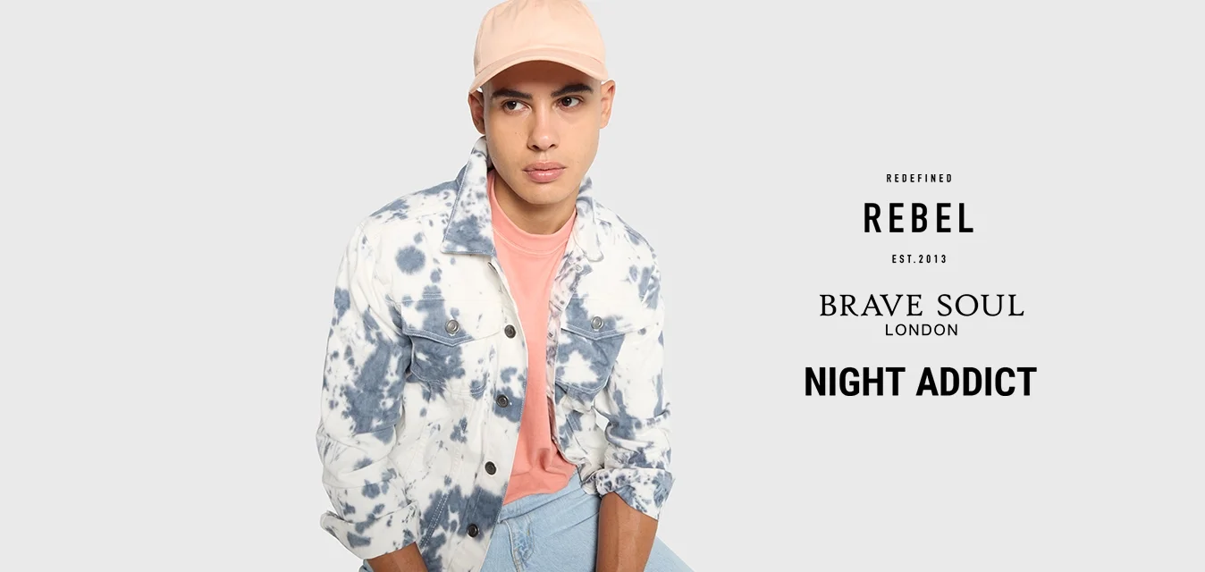 Giovane uomo che indossa un cappello rosa, una camicia corallo e una giacca tie-dye blu e bianca, seduto e che guarda leggermente di lato su uno sfondo grigio.