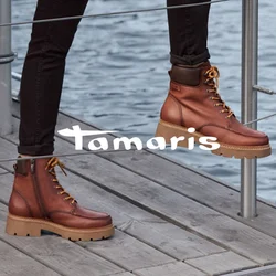 Cappotto impermeabile marrone con colletto, maglione crema, pantaloni neri e stivaletti marroni con lacci e suola spessa. Contesto in legno e metallo.