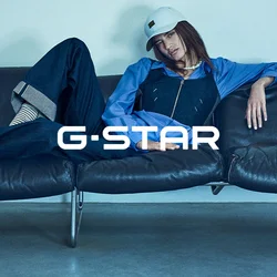 Outfit in denim con una camicia blu e maniche a fantasia, abbinato a un cappellino bianco. Il modello è seduto su un divano in pelle scura, con le gambe incrociate. Calzini a righe visibili.