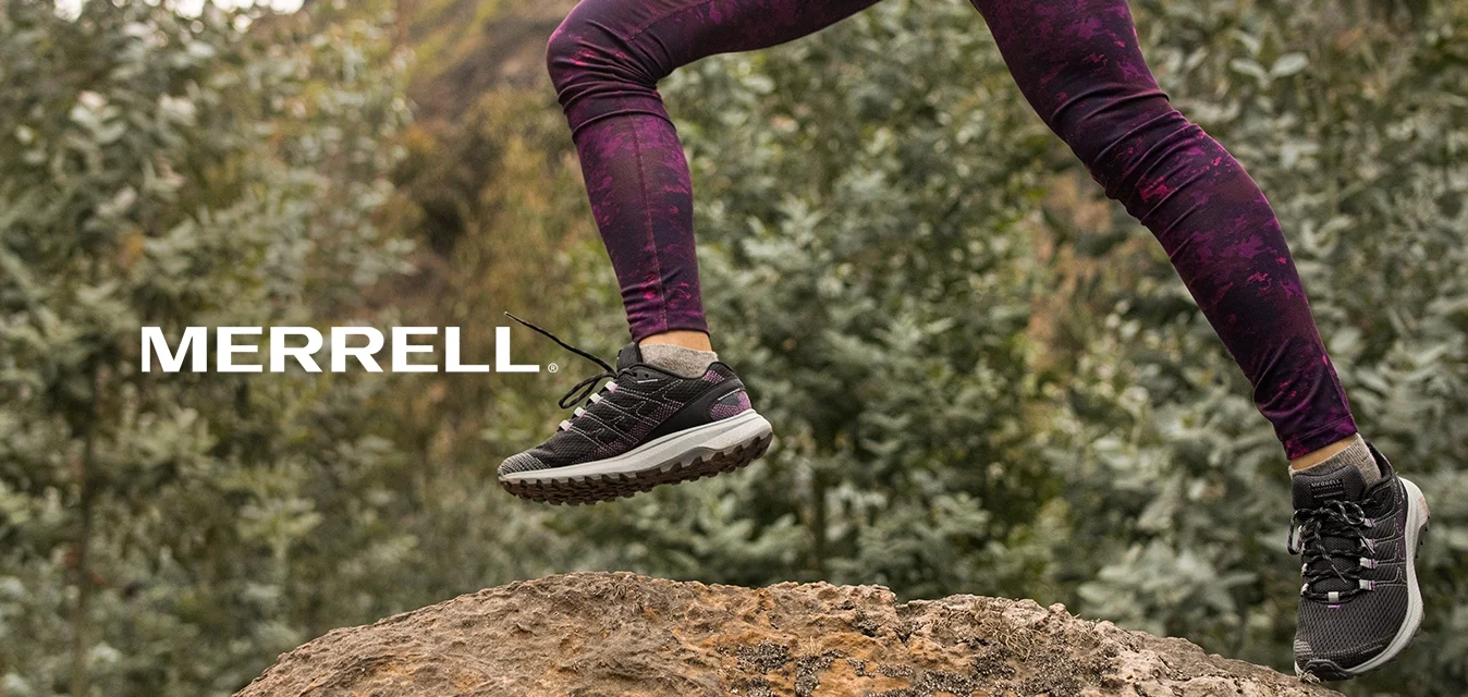 Persona che indossa leggings viola e sneakers neri mentre salta sopra un grande sasso in un'area boschiva all'aperto.